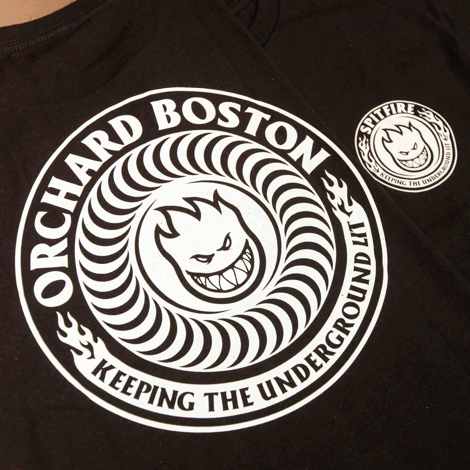 A black t-shirt featuring a large white circular graphic with the text 'ORCHARD BOSTON' and 'KEEPING THE UNDERGROUND LIT' around a cartoon face, plus a smaller matching circular patch on the sleeve.